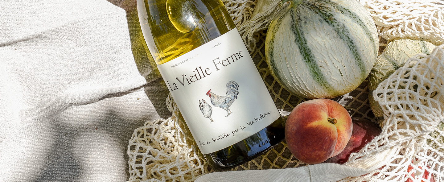 Bottle of La Vieille Ferme white wine resting on lace linen beside a fresh cantaloupe melon and a ripe peach