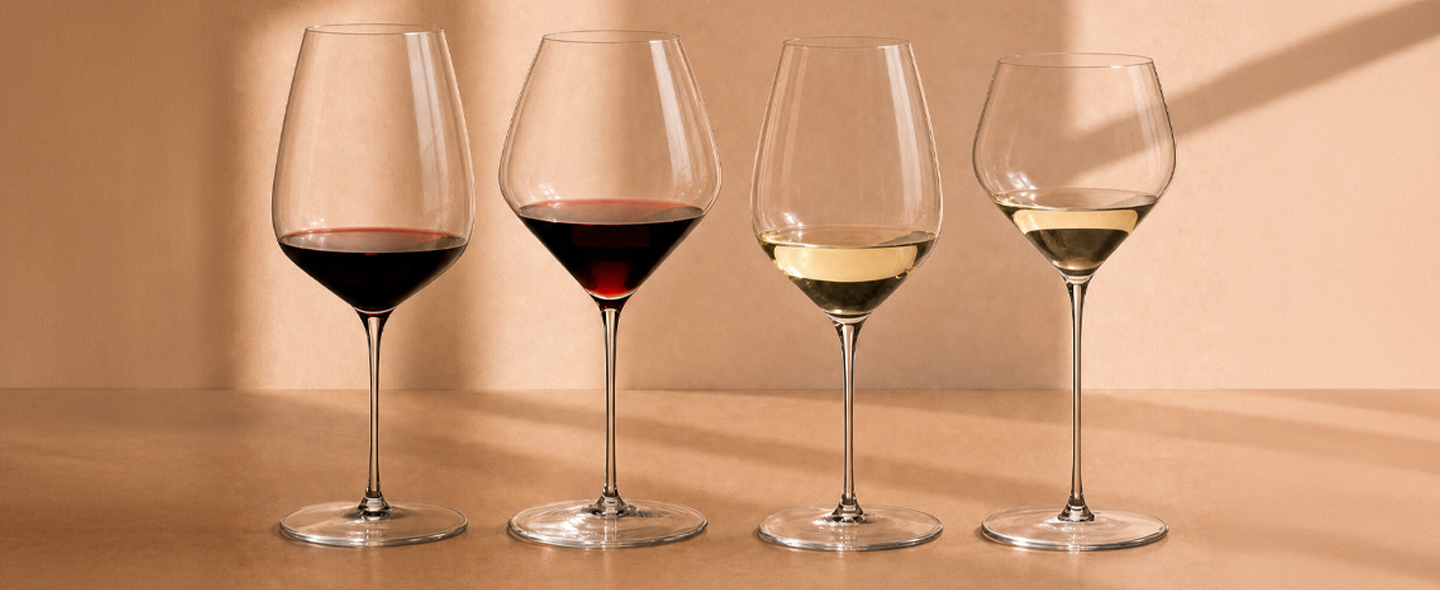 Four Riedel crystal wine glasses in a row — two red wine glasses and two white wine glasses — arranged on a wooden surface against a warm amber background