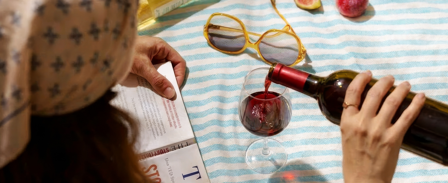 Red wine being poured into a glass beside sunglasses on a striped picnic blanket.