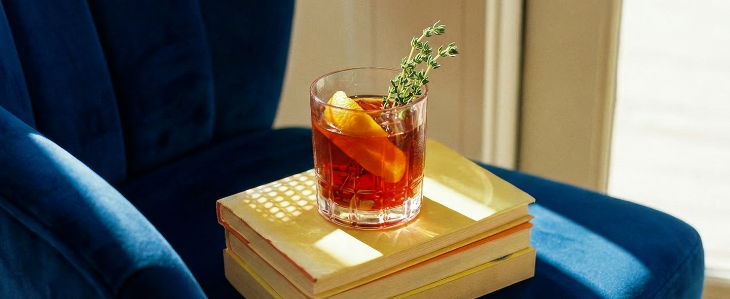Cocktail with orange peel and herbs on books beside a blue velvet armchair.