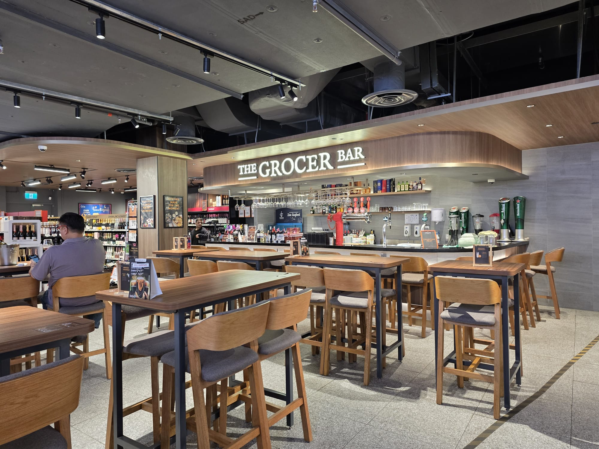The Grocer Bar at FairPrice Finest Sembawang Shopping Centre with high wooden tables and chairs.
