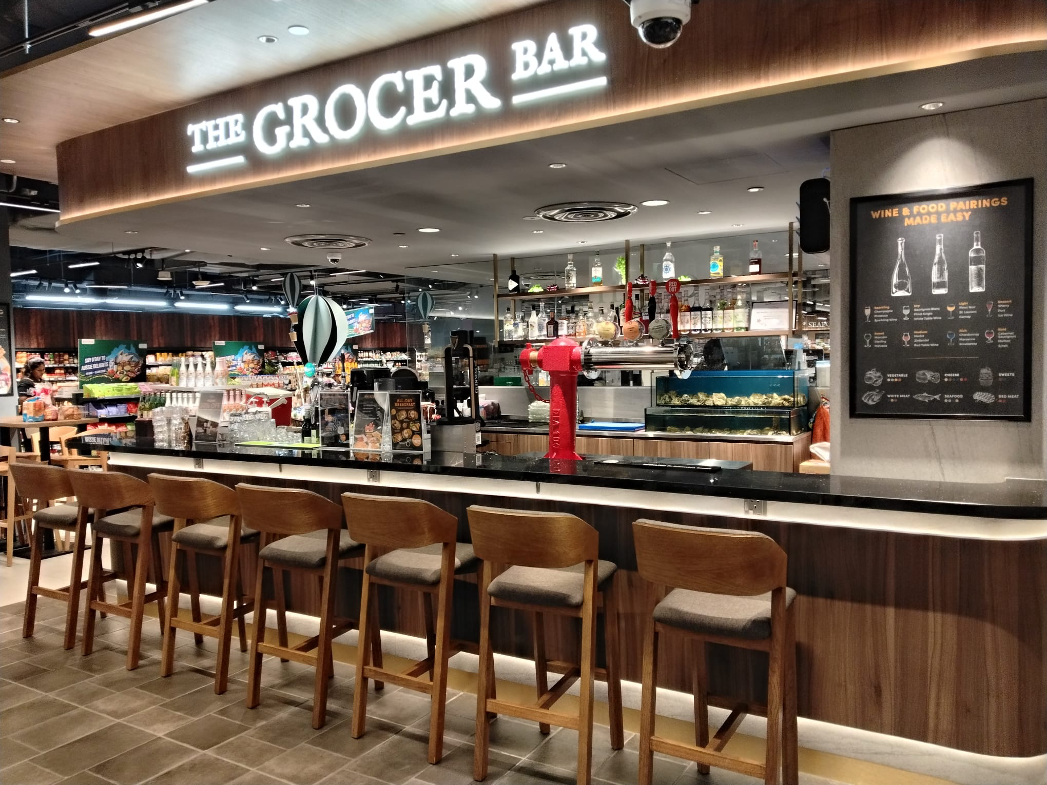 The Grocer Bar at FairPrice Finest Thomson Plaza with wooden barstools, a red beer tap and framed menu.
