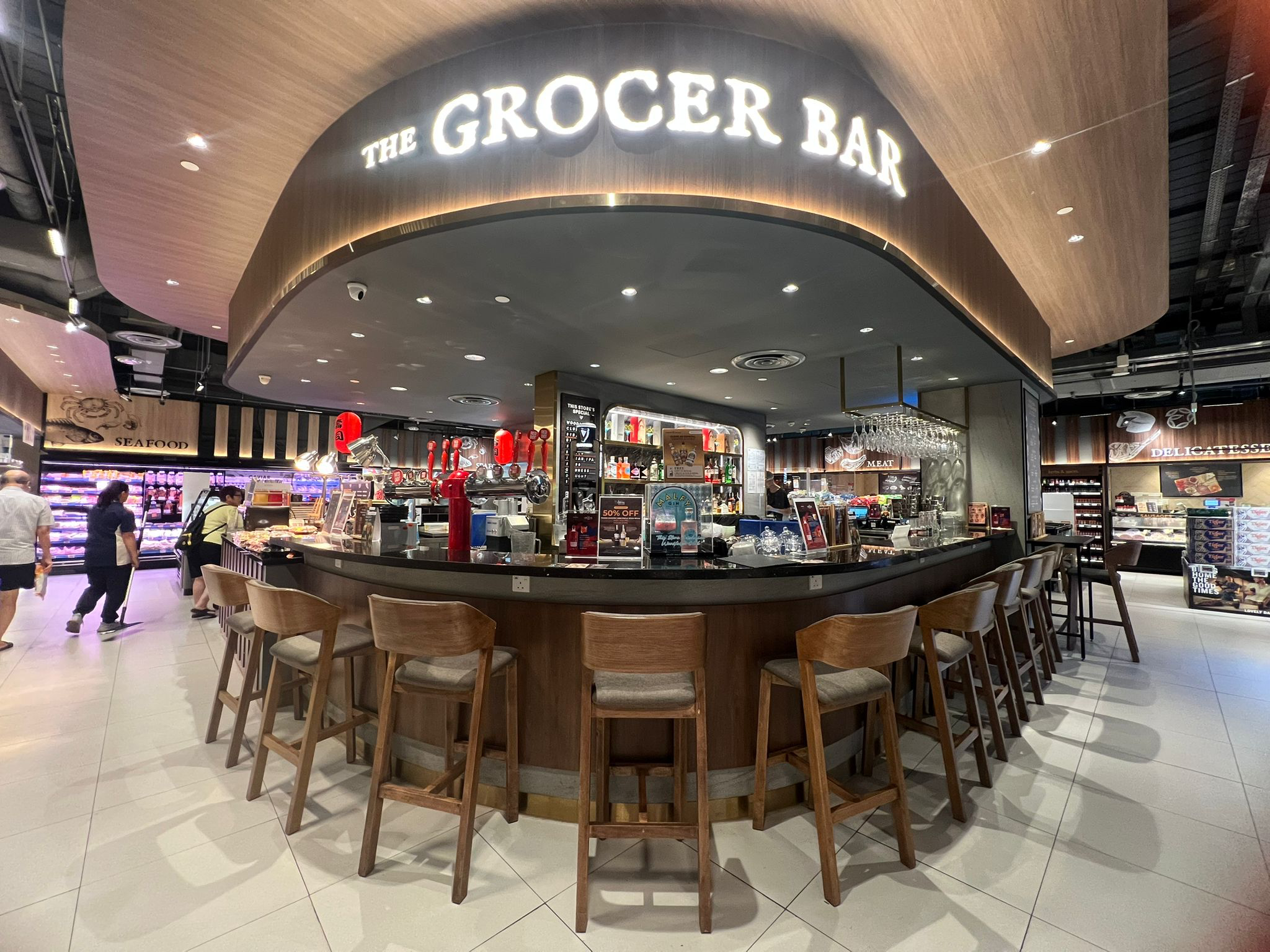 The Grocer Bar at FairPrice Finest Woodleigh Mall with a curved counter and wooden barstools.