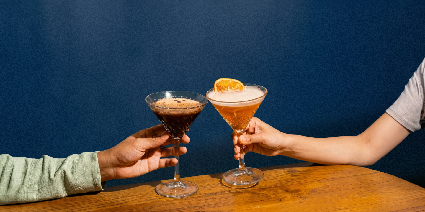 Two cocktail glasses clinking, garnished with orange slices, against a dark blue background.